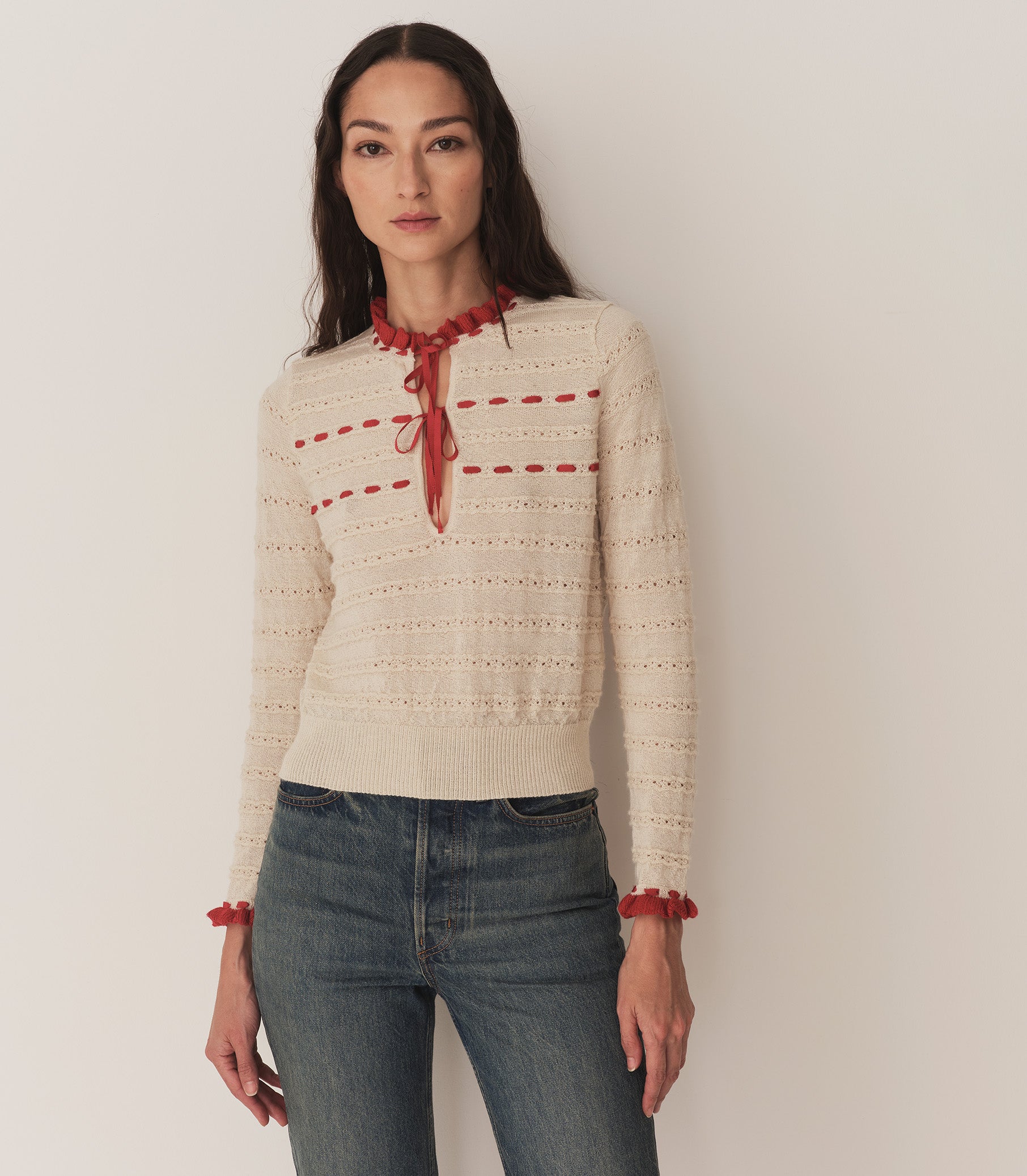 A woman wearing jeans and the Cordelie Top, in a cream and red baby alpaca-silk blend and silk grosgrain ribbons that tie at the neck. 3