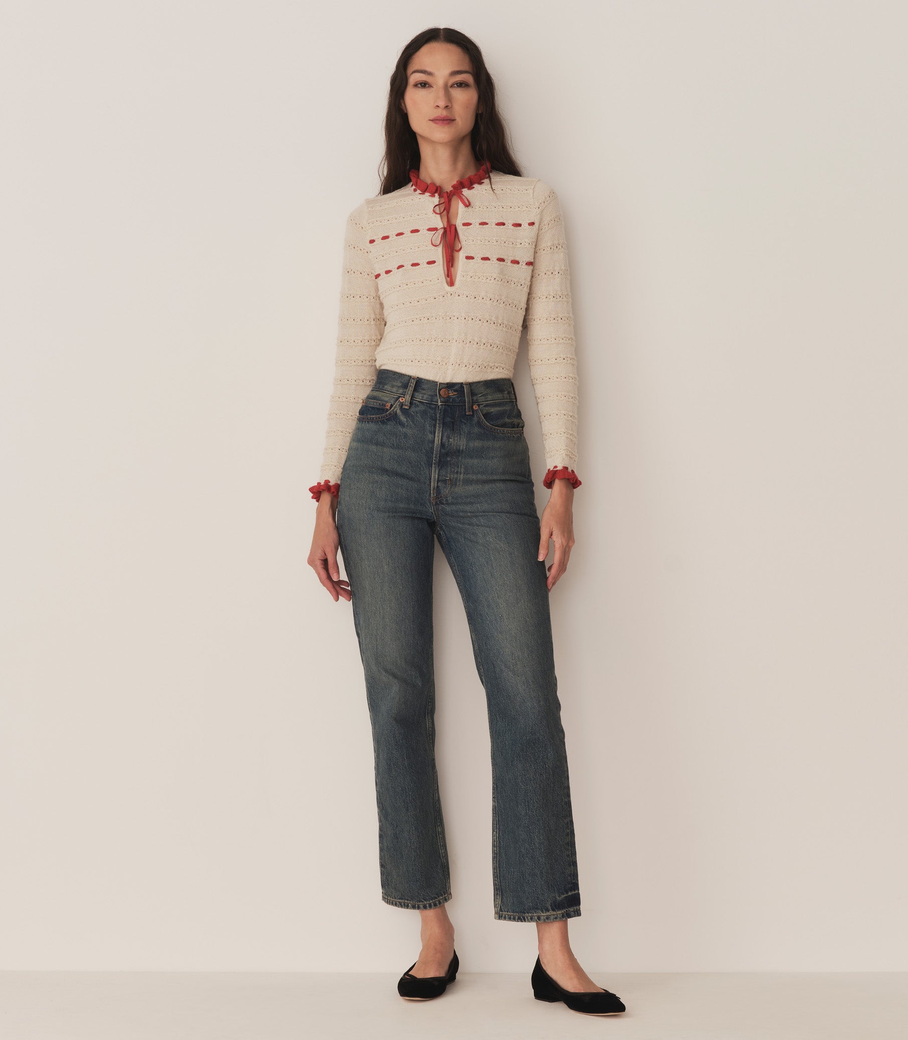 A woman wearing jeans and the Cordelie Top, in a cream and red baby alpaca-silk blend and silk grosgrain ribbons that tie at the neck. 2