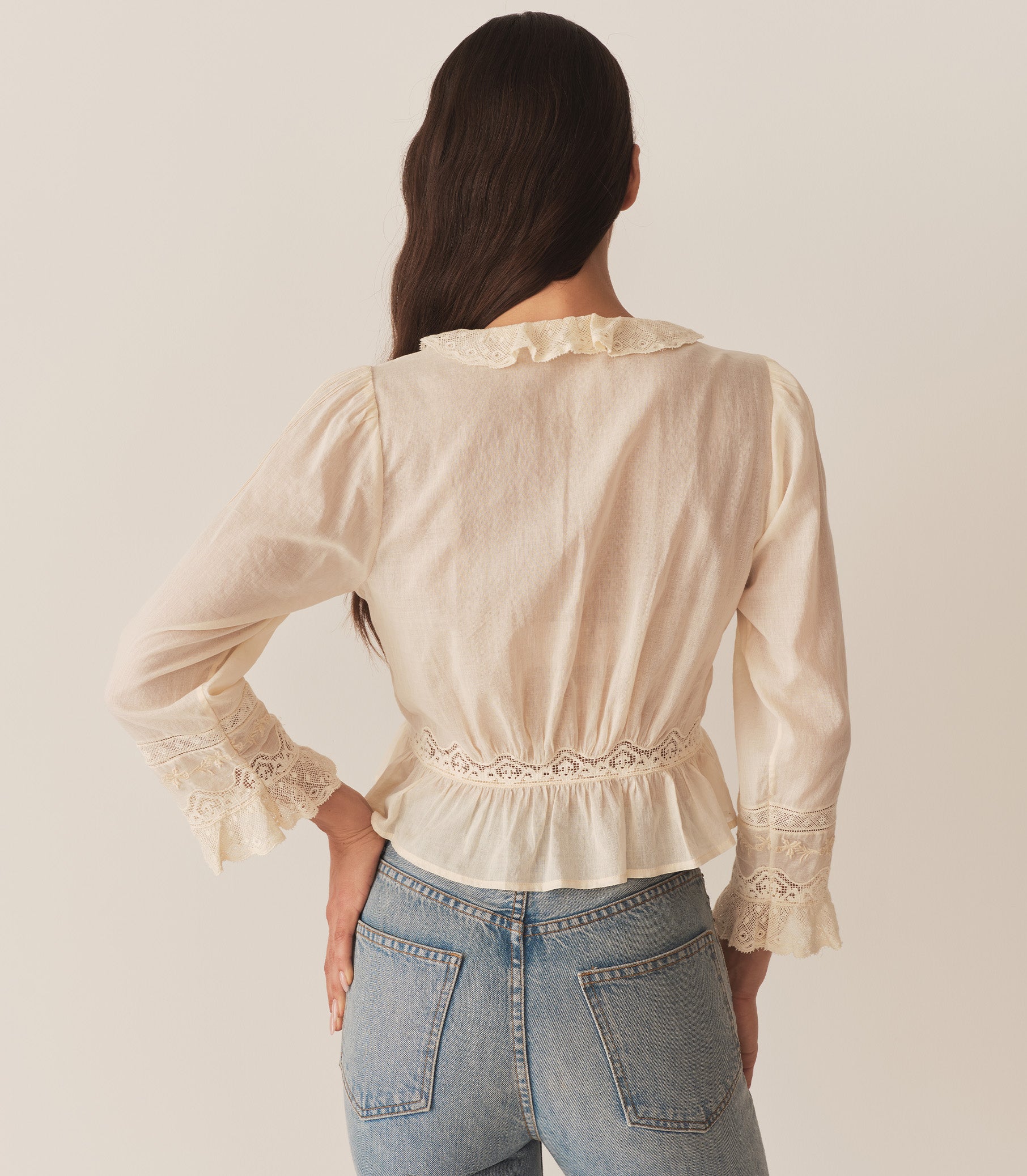 A woman wearing the Eldoris Top in Alabaster, made in organic cotton voile and featuring a plunging V-neckline framed with cascading lace-trimmed ruffles that extend down the bodice and alternating panels of inset lace, clustered pintucks, and intricate embroidery at the yoke and back. 4