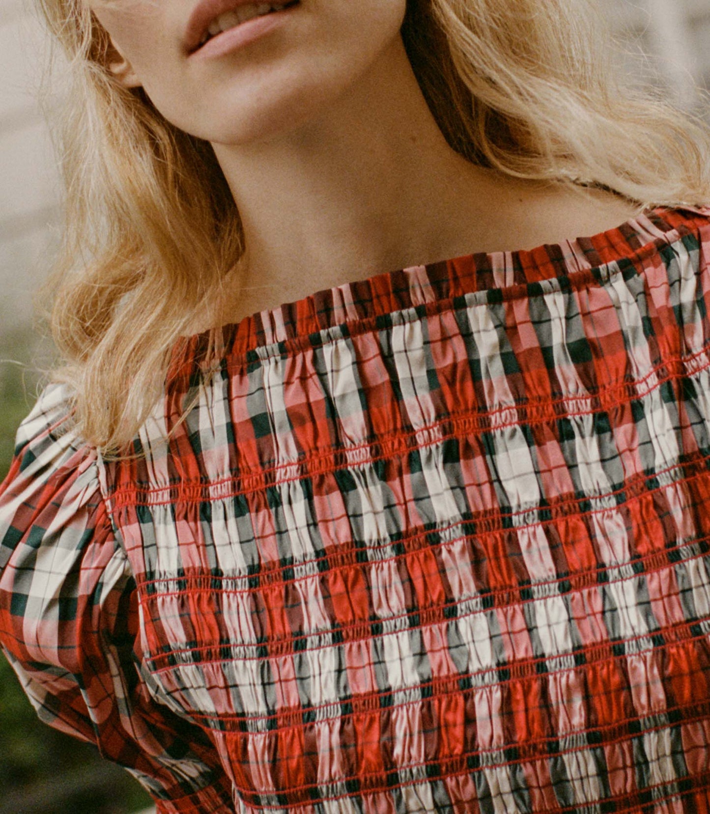 A woman wearing the Raquella Dress in Rouge Adaline Plaid, a red-and-white plaid dress with smocking at the bust and puffed sleeves, with a black belt tied at her waist.